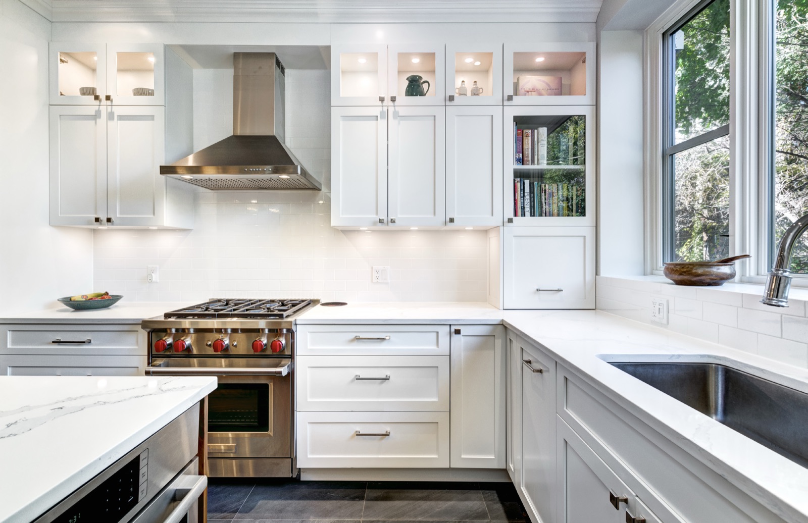 A kitchen with White Shaker cabinets, glass-front wall cabinets and a stainless range hood